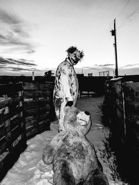 A sinister clown with a large teddy bear between wooden haunt walls